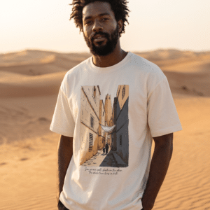 Man wearing the beige Ghardaïa tee in the Sahara desert, printed with an illustrated alleyway from Algeria's M'Zab Valley