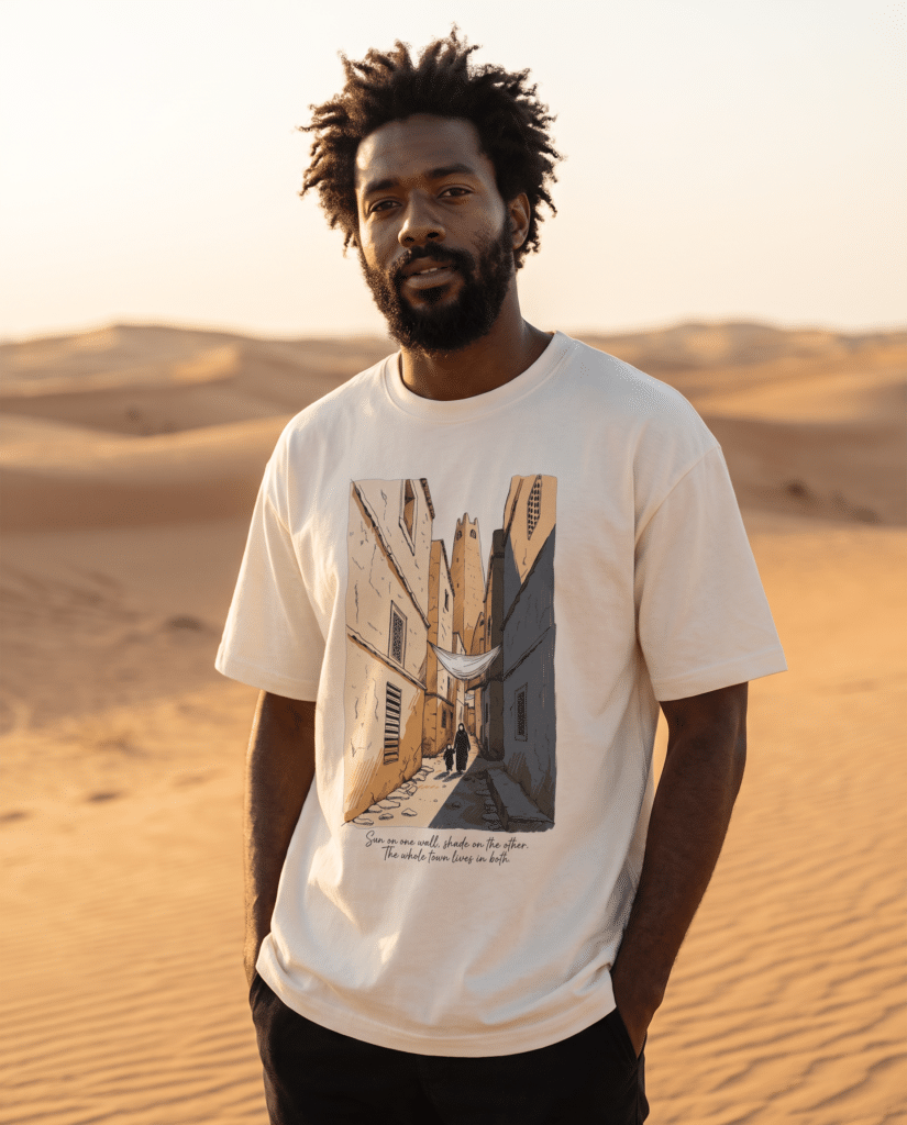 Man wearing the beige Ghardaïa tee in the Sahara desert, printed with an illustrated alleyway from Algeria's M'Zab Valley